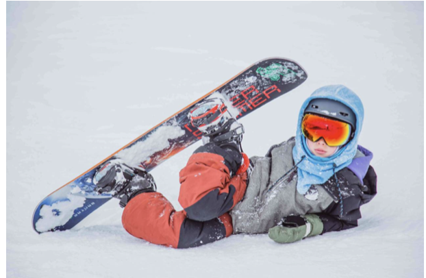A child in a helmet and snowsuit lying on the snow beside a snowboard